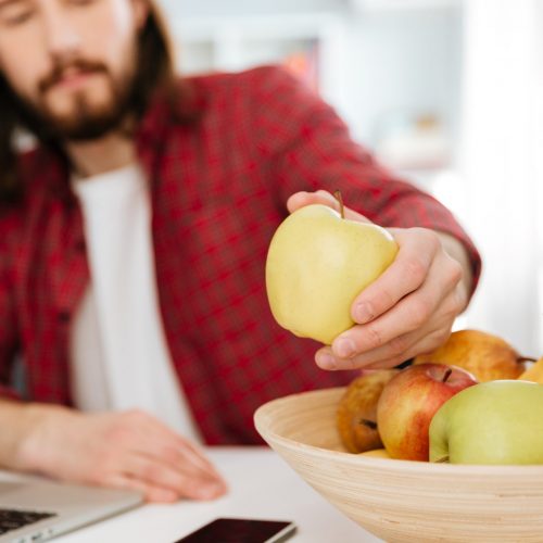 Closeup of bearded young man eating fruits and using laptop at home