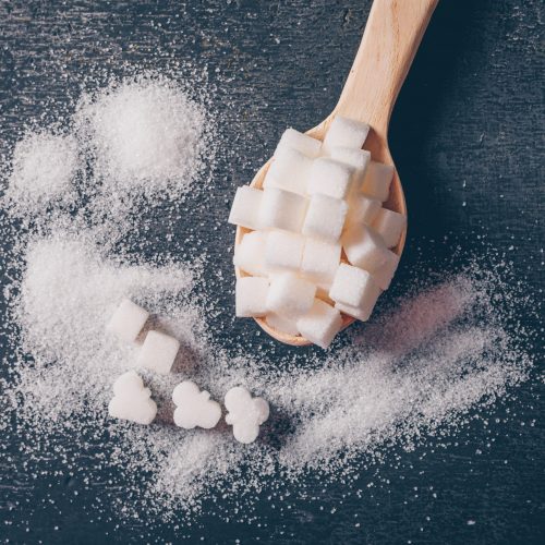 Some white sugar in a spoon on dark background, flat lay.